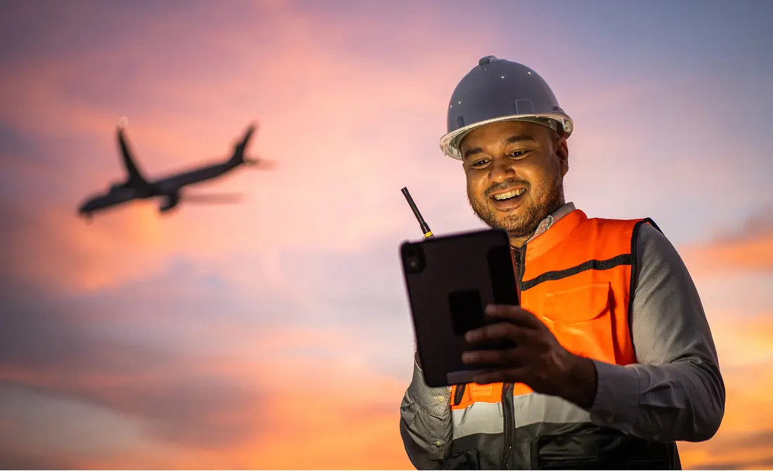 Colaborador de OMA con casco y chaleco de seguridad consulta una tableta en el aeropuerto mientras un avión despega al fondo